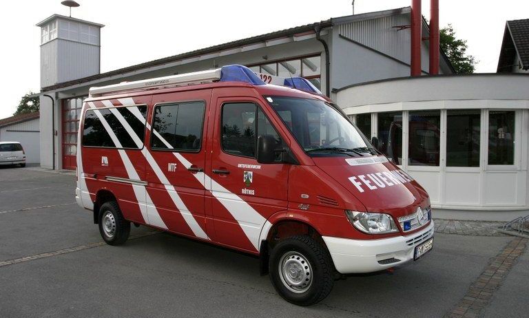 Ein rot-weißer Feuerwehrwagen mit der Aufschrift 'FEUERWEHR' vorne ist vor einer Feuerwache geparkt. Der Wagen hat ein blaues Dach mit blinkenden Lichtern. Die Feuerwache ist weiß und hat einen Turm mit Glasfenstern.