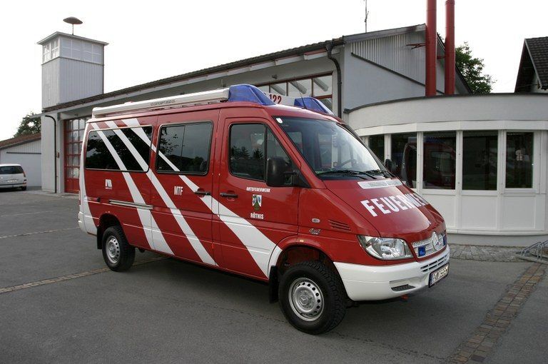 Ein rot-weißer Feuerwehrwagen mit der Aufschrift 'FEUERWEHR' vorne ist vor einer Feuerwache geparkt. Der Wagen hat ein blaues Dach mit blinkenden Lichtern. Die Feuerwache ist weiß und hat einen Turm mit Glasfenstern.