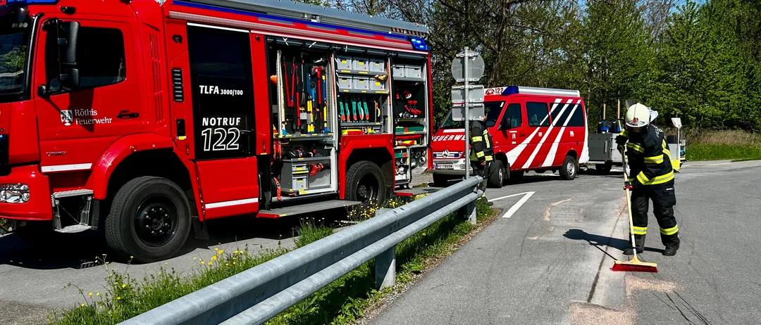 Ein roter Feuerwehrwagen mit der Aufschrift NOTRUF 122 und ein roter Rettungswagen sind am Straßenrand geparkt. Ein Feuerwehrmann fegt die Straße.