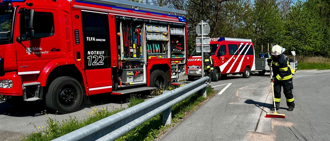 Ein roter Feuerwehrwagen mit der Aufschrift NOTRUF 122 und ein roter Rettungswagen sind am Straßenrand geparkt. Ein Feuerwehrmann fegt die Straße.
