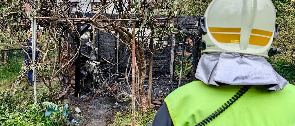 Ein Feuerwehrmann mit den Initialen TLF steht vor einem ausgebrannten Schuppen, neben einem Gartenschlauch und einem verbrannten Baum.