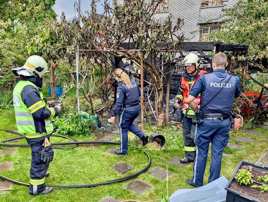Feuerwehrleute und Polizei inspizieren einen verbrannten Garten. Ein Feuerwehrmann hält ein Werkzeug, ein anderer geht vorbei. Ein Polizeibeamter steht rechts und beobachtet.