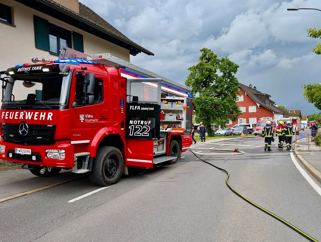 Ein roter Feuerwehrwagen mit der Nummer 122 steht am Straßenrand. In der Nähe sind zwei Feuerwehrleute, und eine Feuerwehrschläuche liegt auf der Straße.