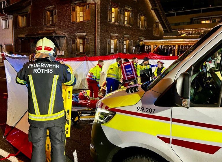 Rettungskräfte in gelben Uniformen arbeiten an einem Patienten in der Nähe eines Krankenwagens. Das Gebäude im Hintergrund hat viele Fenster und festliche Lichter.