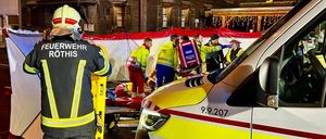 Rettungskräfte in gelben Uniformen arbeiten an einem Patienten in der Nähe eines Krankenwagens. Das Gebäude im Hintergrund hat viele Fenster und festliche Lichter.
