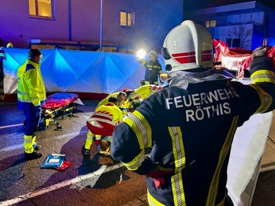 Feuerwehrleute in Schutzkleidung kümmern sich nachts um eine verletzte Person auf der Straße. Ein Rettungswagen und ein Gebäude sind im Hintergrund zu sehen.