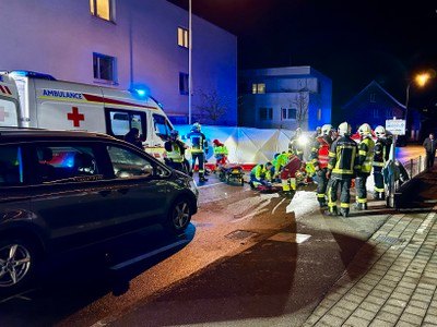 Einsatzkräfte, darunter Feuerwehrleute, kümmern sich um verletzte Personen am Unfallort auf einer Straße bei Nacht. Ein Krankenwagen ist in der Nähe geparkt, und ein Auto ist hochgezogen.