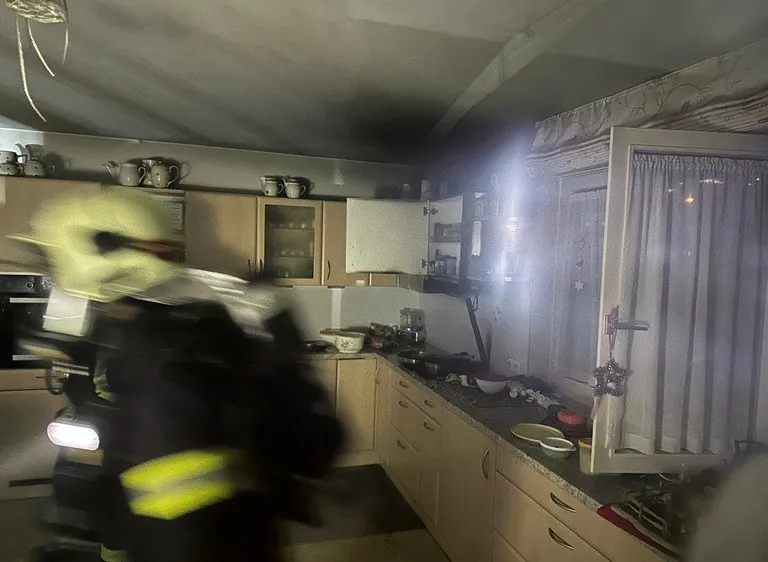 Ein Feuerwehrmann ist in einer Küche, die zu brennen scheint. Die Küche ist schwach beleuchtet, und Rauch steigt aus dem Bereich auf.