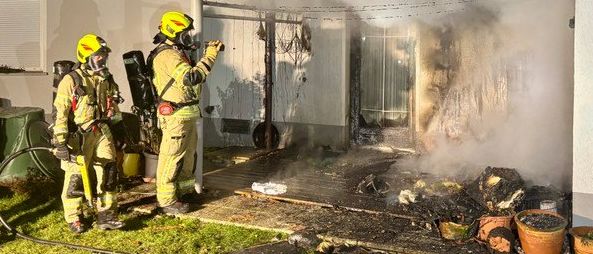 Feuerwehrleute in Schutzkleidung arbeiten daran, einen Hausbrand zu löschen. Rauch und Flammen entweichen aus dem Dach und dem Erdgeschoss.