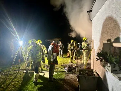 Feuerwehrleute in Schutzkleidung stehen bei Nacht vor einem Gebäude, aus dem Rauch aufsteigt. Ein Mann in einem roten Schal steht unter ihnen, und sie werden von einem hellen Licht beleuchtet.