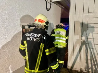 Zwei Feuerwehrleute, einer in einer Rothis-Feuerwehruniform und der andere in einer gelben Sicherheitsweste, stehen neben einer offenen Tür, die von einem Licht beleuchtet wird. Ein Schatten und ein Stativ sind an der Wand zu sehen.