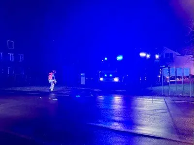 Ein Feuerwehrmann steht auf einer nassen Straße bei Nacht, beleuchtet von blauen Lichtern der Einsatzfahrzeuge. Im Hintergrund sind Häuser zu sehen, und ein Auto ist vor einem geparkt.