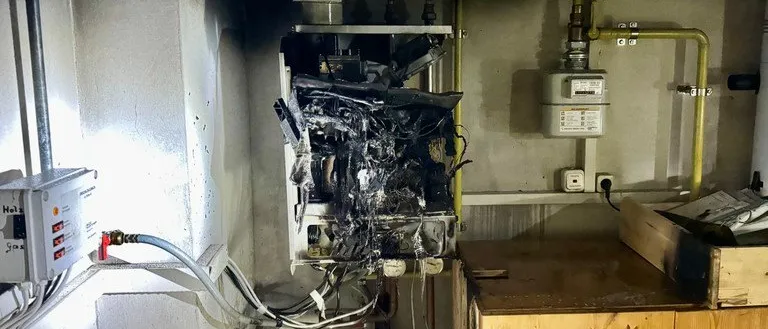 Ein verbranntes und beschädigtes Gerät ist an einer Wand montiert, mit daran angeschlossenen Kabeln und Rohren. Ein hölzerner Schrank ist auf der rechten Seite platziert, und ein Gaszähler ist über dem Schrank an der Wand montiert.