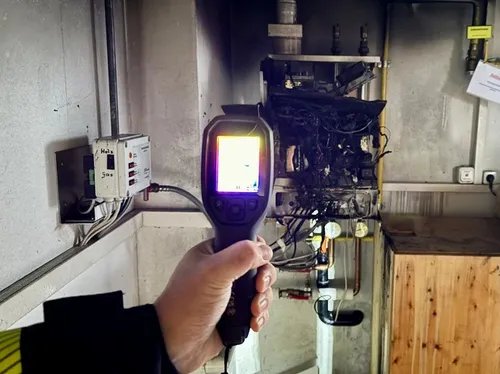 Eine Hand, die ein Gerät hält, ist an einer Wand mit elektrischen Geräten und Rohren zu sehen. Das Gerät zeigt eine Anzeige.