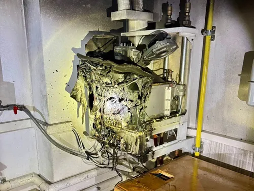 Ein beschädigtes elektrisches Gehäuse, bedeckt mit Ruß und Kabeln, liegt an einer Wand. Das Gehäuse weist sichtbare Brandflecken auf und ist an einen Rohr und einen gelben Schlauch angeschlossen.