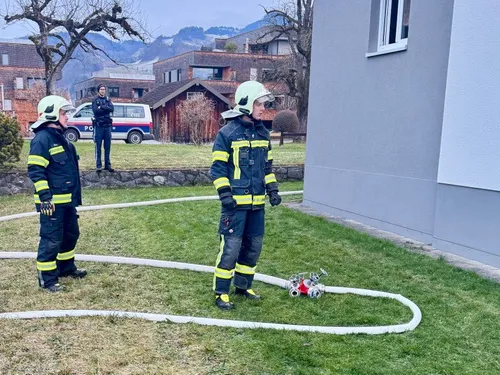 Zwei Feuerwehrleute in Uniform stehen neben einem Gebäude mit einem Hydranten auf dem Rasen. Ein Polizeibeamter steht hinter ihnen. Im Hintergrund befinden sich Häuser, Bäume und ein Van.
