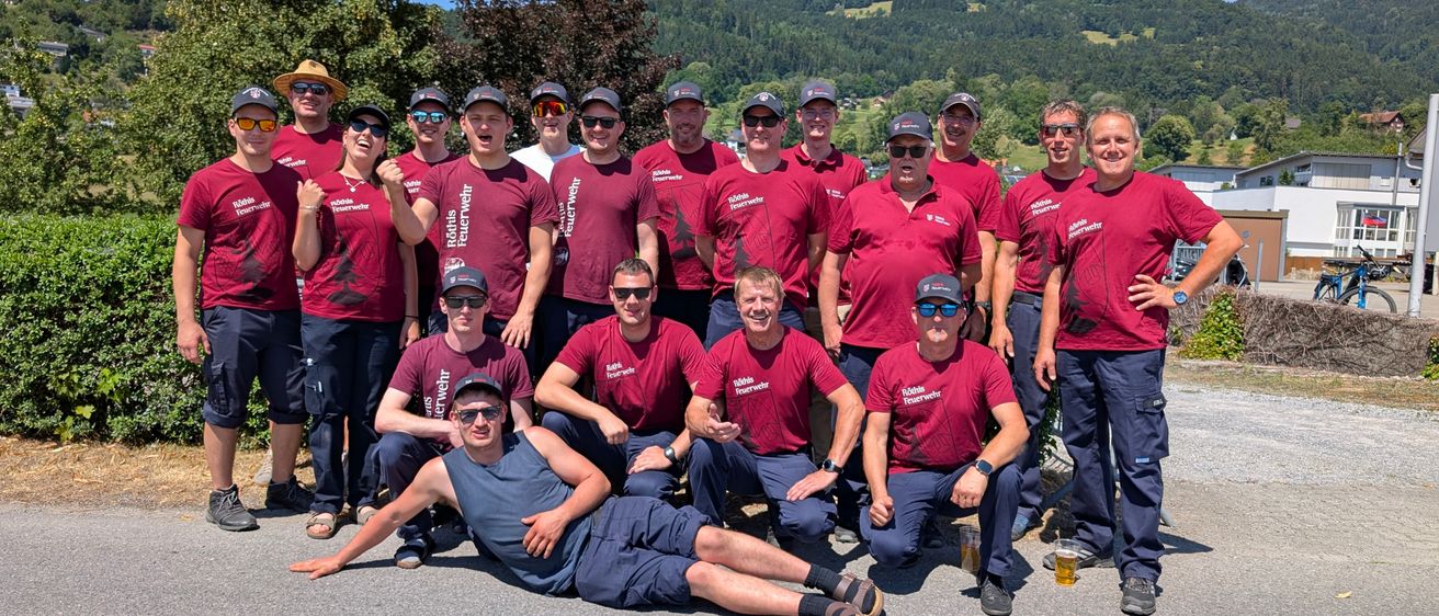 Eine Gruppe von Menschen in roten T-Shirts und Mützen posiert für ein Foto mit Bergen im Hintergrund. Einige sitzen, andere stehen.