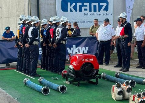 Eine Gruppe Feuerwehrleute in Uniform steht in einer Reihe mit Helmen auf dem Kopf, einem roten und schwarzen Feuerwehrroboter vor ihnen und einem Banner mit dem Text 'kranz luft-klima-technik' im Hintergrund.