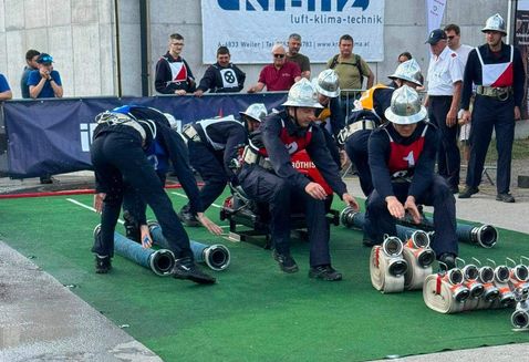 Eine Gruppe von Feuerwehrleuten in Helmen und Uniformen bereitet sich auf ein Rennen auf einem grünen Feld vor, einige beugen sich und andere sitzen auf Karren. Zuschauer beobachten hinter einem Zaun.