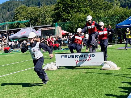 Eine Gruppe junger Menschen in roten und weißen Uniformen nimmt an einer Jugendveranstaltung der Feuerwehr teil, läuft und springt über Hindernisse auf einem Feld.