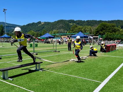 Eine Gruppe von Menschen in gelben Westen nimmt an einer Sportveranstaltung auf einem Feld mit Kunstrasen teil. Eine Person springt über eine Hürde, während andere sich auf den Lauf vorbereiten.
