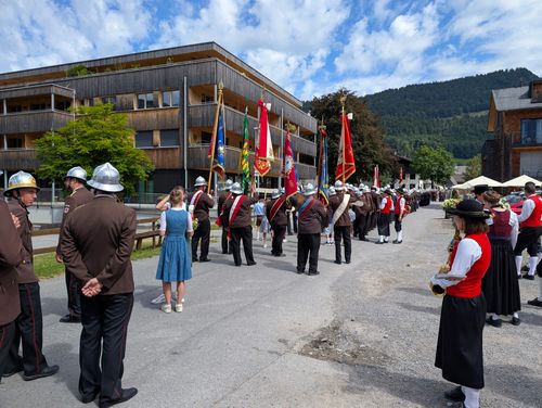 Eine Marschkapelle in traditionellen Uniformen und Helmen steht in Formation auf einer Straße mit Gebäuden und Bergen im Hintergrund.