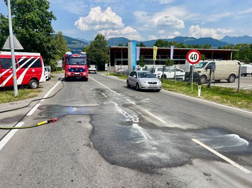 Ein Feuerwehrwagen fährt auf einer Straße mit einem Geschwindigkeitsbegrenzungsschild von 40 km/h. Ein Auto fährt hinter ihm. Es gibt eine Pfütze auf der Straße.