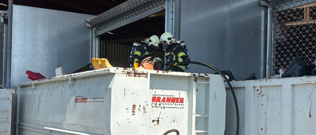 Zwei Feuerwehrleute in Schutzkleidung stehen neben einem großen weißen Müllcontainer mit einer Leiter. Der Container ist Teil einer Reihe ähnlicher Behälter.