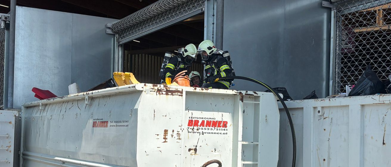 Zwei Feuerwehrleute in Schutzkleidung stehen neben einem großen weißen Müllcontainer mit einer Leiter. Der Container ist Teil einer Reihe ähnlicher Behälter.