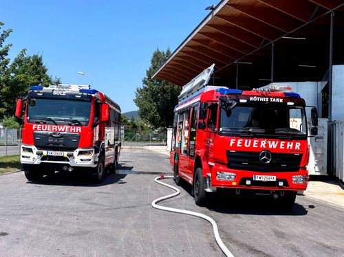 Zwei rote Feuerwehrfahrzeuge sind in einer Feuerwache geparkt, eines mit dem Wort 'Feuerwehr' an der Seite. Eine Feuerwehrschläuche ist an einem der Fahrzeuge angeschlossen.