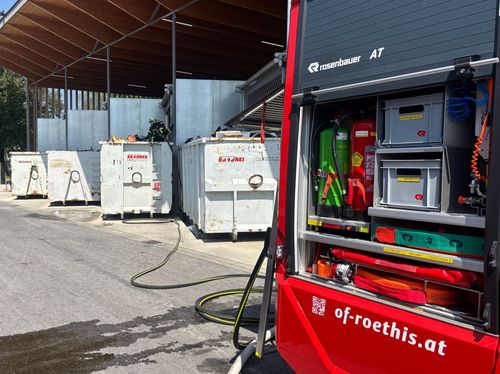 Ein Feuerwehrauto steht in einer Garage mit offener Hecktür, wobei Feuerlöscher und Ausrüstung im Inneren zu sehen sind. Ein Feuerwehrmann steht in der Nähe des Fahrzeugs. Mehrere weiße Container sind hinter dem Fahrzeug aufgereiht.