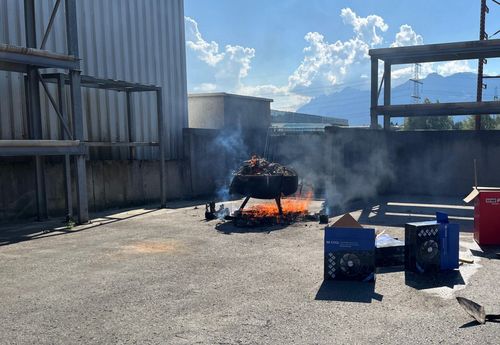 Ein Lagerfeuer brennt in einem Außenbereich, umgeben von Industriegebäuden, wobei Rauch in den Himmel steigt. Ein Karton steht in der Nähe auf dem Boden.
