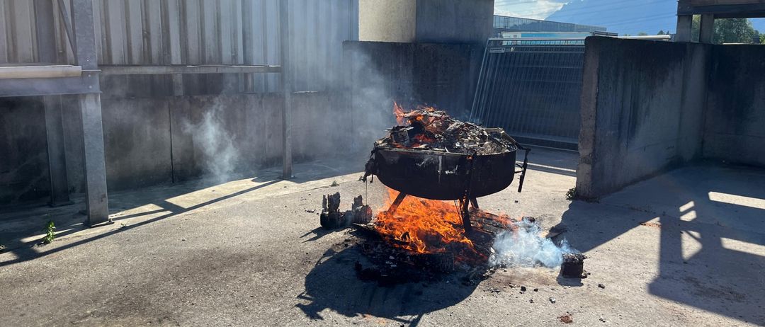 Ein Feuerkorb auf einer Betonfläche im Freien mit aufsteigendem Rauch, umgeben von Betonstrukturen und einem klaren blauen Himmel.