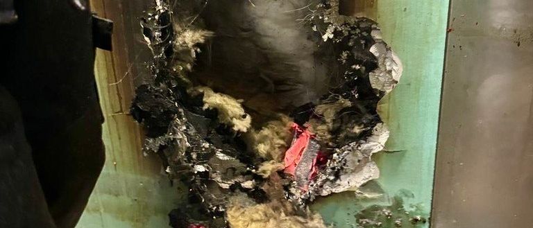 Eine Nahaufnahme einer beschädigten Wand mit einem Loch und verkohlten Materialien, möglicherweise von einem Feuer. Die Wand ist grün und hat eine metallische Textur.