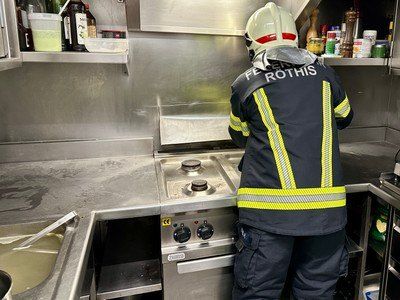 Ein Feuerwehrmann in einer Küche, der in der Nähe eines Herdes mit dem Namen 'Feuer Rothis' auf seiner Uniform steht und den Bereich inspiziert.