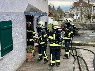 Eine Gruppe von Feuerwehrleuten in voller Ausrüstung, einschließlich Helmen und Masken, steht auf einer Holzterrasse vor einem Gebäude. Ein Feuerwehrmann betritt das Gebäude durch eine Tür.