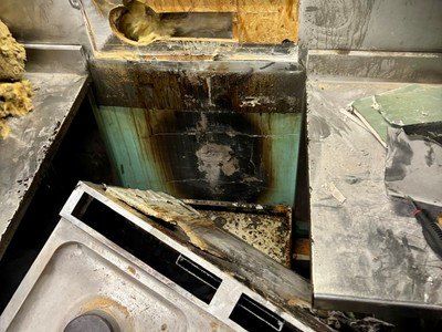 Ein beschädigter Küchenbereich mit einem verbrannten und rostigen Waschbecken, einem kaputten Gerät und verstreutem Schutt. Die Wände sind mit grünen Fliesen verkleidet, von denen einige beschädigt sind.