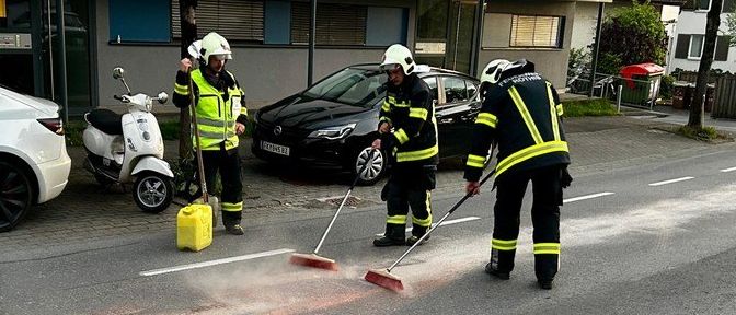 Drei Feuerwehrleute in gelben Westen reinigen die Straße mit Besen. Ein weißes Auto und ein Roller sind auf der Seite geparkt.