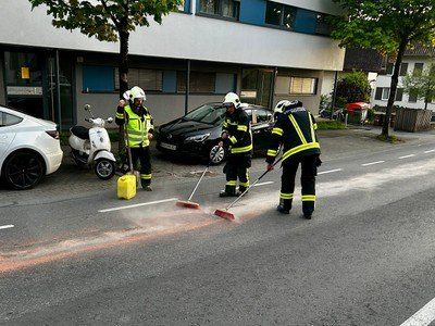Drei Feuerwehrleute reinigen eine Straße mit Besen. Ein weißes Auto, ein Roller und ein schwarzes Auto sind in der Nähe geparkt. Ein Gebäude mit blauen Verkleidungen ist im Hintergrund.