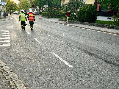 Drei Personen gehen auf einer Straße mit weißen Markierungen; zwei tragen reflektierende Westen und eine ist in Freizeitkleidung. Die Straße ist leer, und es gibt Gebäude und Grünflächen an den Seiten.