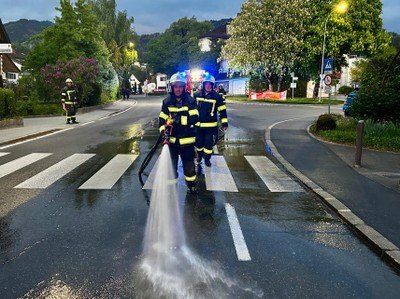 Drei Feuerwehrleute reinigen eine nasse Straße mit einem Schlauch, einer sprüht Wasser auf einen Zebrastreifen. Bäume und Straßenlaternen sind im Hintergrund zu sehen.