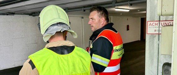 Zwei Feuerwehrleute stehen in einer schwach beleuchteten Garage. Einer trägt eine gelbe Weste mit den Buchstaben AFK, der andere eine orange Weste mit dem Wort Einsat.