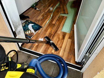 Eine Person benutzt einen Staubsauger, um einen Holzboden in einem Haus zu reinigen. Der Boden scheint einer Renovierung zu unterliegen, wobei einige Holzbretter entfernt und eine grüne Matte platziert wurden.