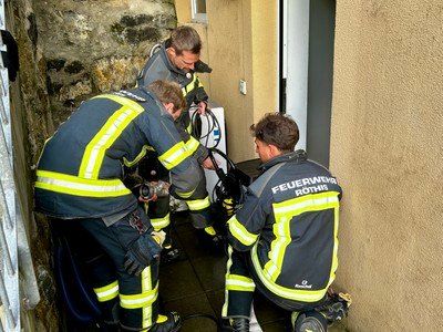 Vier Feuerwehrleute in Schutzkleidung arbeiten in einem engen Raum, möglicherweise am Eingang eines Hauses. Einer hält einen Schlauch, ein anderer kniet, und zwei andere stehen.