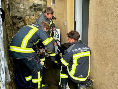 Vier Feuerwehrleute in Schutzkleidung arbeiten in einem engen Raum, möglicherweise am Eingang eines Hauses. Einer hält einen Schlauch, ein anderer kniet, und zwei andere stehen.