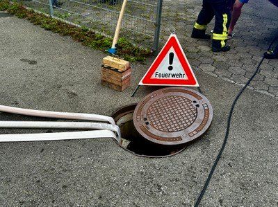 Ein Feuerlöscher mit Warnzeichen und offenem Kanaldeckel auf einem gepflasterten Bereich mit einem Besen in der Nähe.