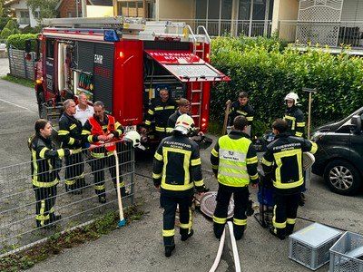 Eine Gruppe von Feuerwehrleuten in gelben und schwarzen Uniformen versammelt sich um einen roten Feuerwehrwagen, einige stehen bei einem Zaun und andere halten Werkzeuge.