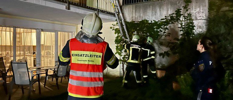 Feuerwehrleute in Schutzkleidung stehen nachts auf einem Balkon, einer klettert eine Leiter. Ein Polizist steht in der Nähe, und ein Mann schaut von einem anderen Balkon herunter.