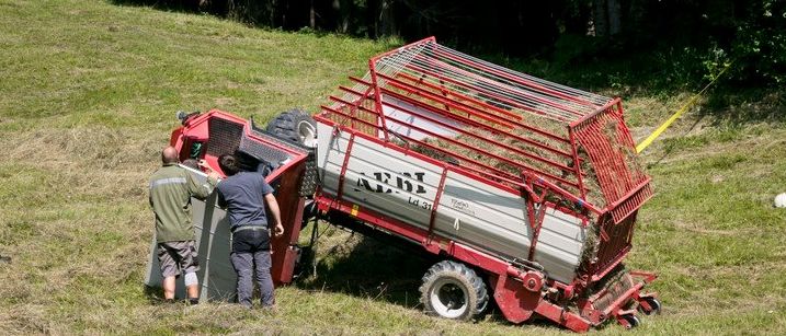 Zwei Männer stehen neben einem umgekippten roten und silbernen landwirtschaftlichen Fahrzeug auf einem Grasfeld.