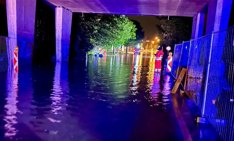 Eine überflutete Straße bei Nacht mit Notfallpersonal in roten Uniformen in der Nähe einer Brücke.